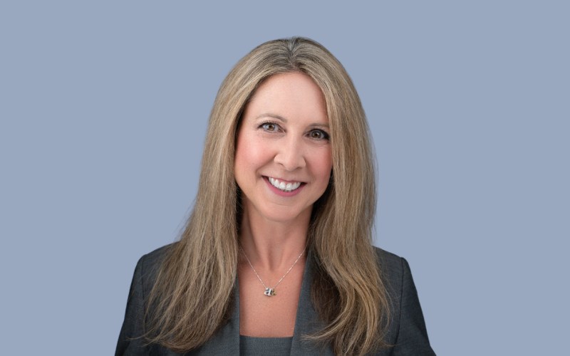 A woman with long, straight blonde hair smiling, wearing a dark blazer and a necklace, standing against a light blue background.