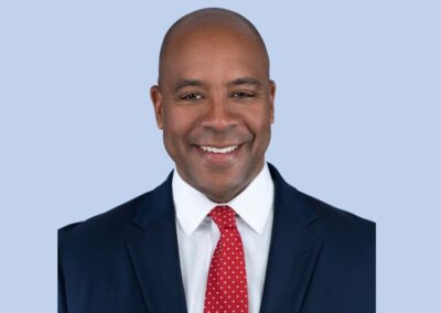 A person in a dark blue suit, white shirt, and red polka dot tie, smiling against a light blue background.
